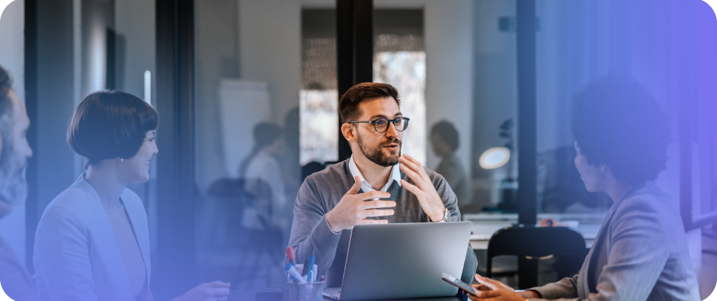 B2B tone of voice A group of people meeting in a modern office. One person speaks while gesturing, another holds a smartphone, and an open laptop and scattered office supplies sit on the table. The background features glass walls and a whiteboard.