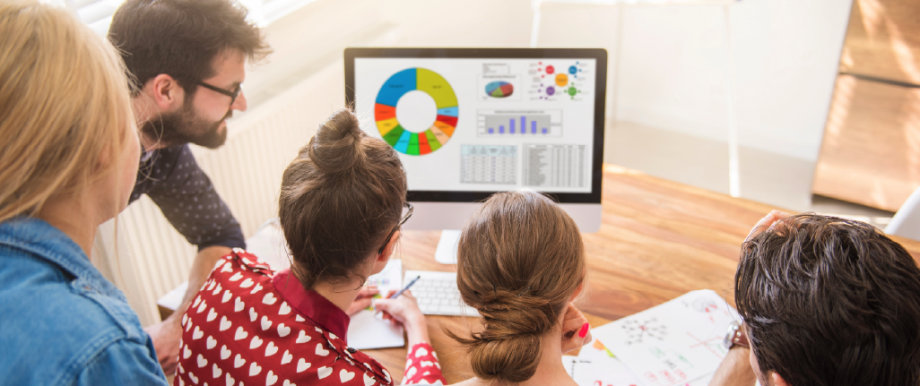 Five individuals are gathered around a computer monitor in a well-lit, modern office setting. The screen displays vibrant data visualizations, including pie charts, bar graphs, and line graphs—suggesting a focus on analytics or strategic planning. The group appears engaged in discussion, with some pointing at the screen and others referencing handwritten notes and diagrams spread across the table. The atmosphere conveys active teamwork, data-driven decision-making, and a blend of digital and analog tools in a professional environment.